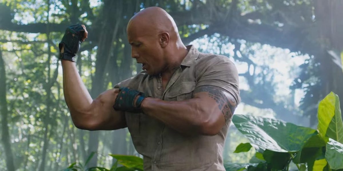 Dr. Xander Bravestone (The Rock) looks shocked at the size of his bicep as he flexes it in 'Jumanji: